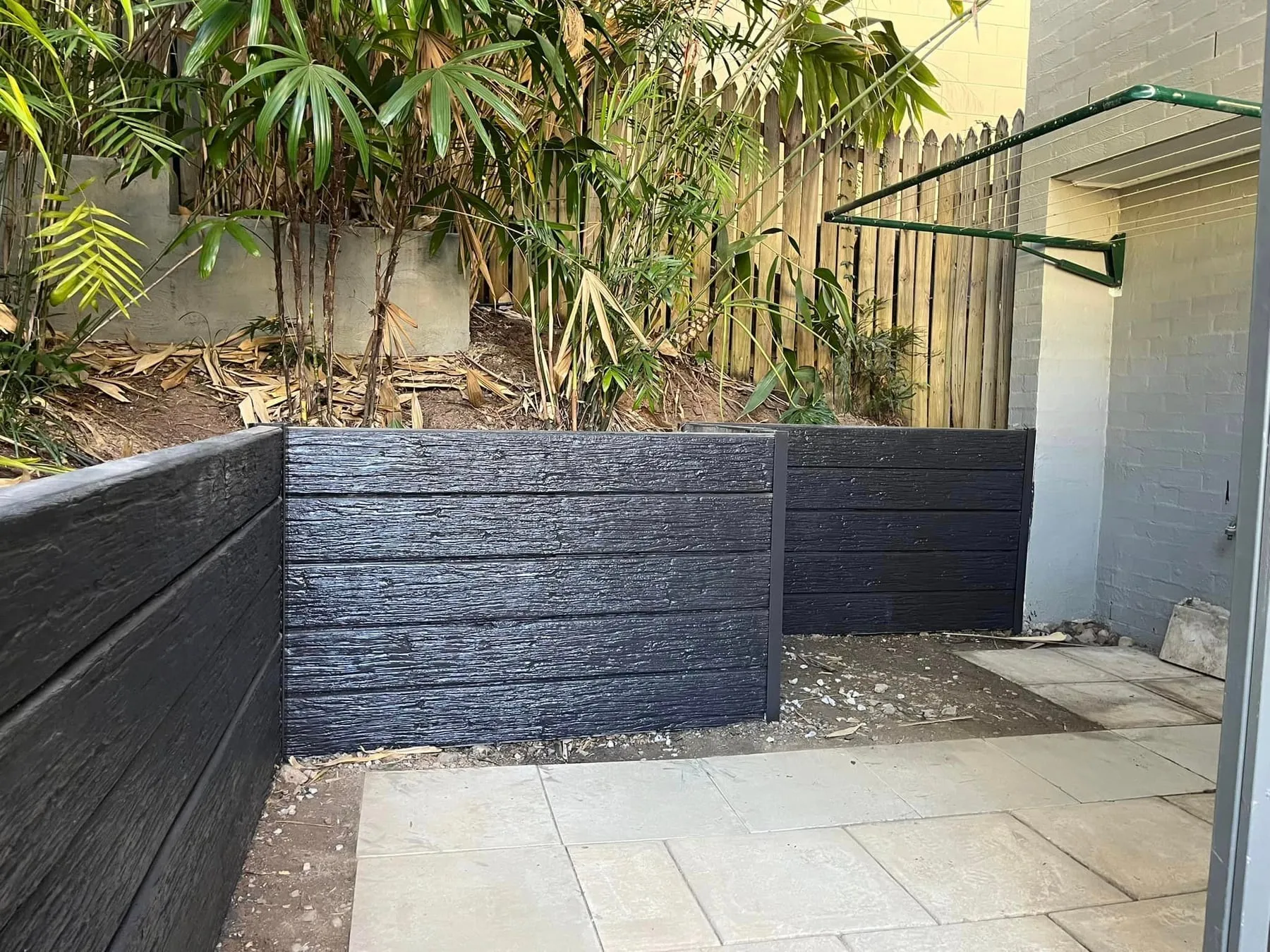 Charcoal woodgrain concrete sleeper retaining wall beside sandstone paving and a palm garden